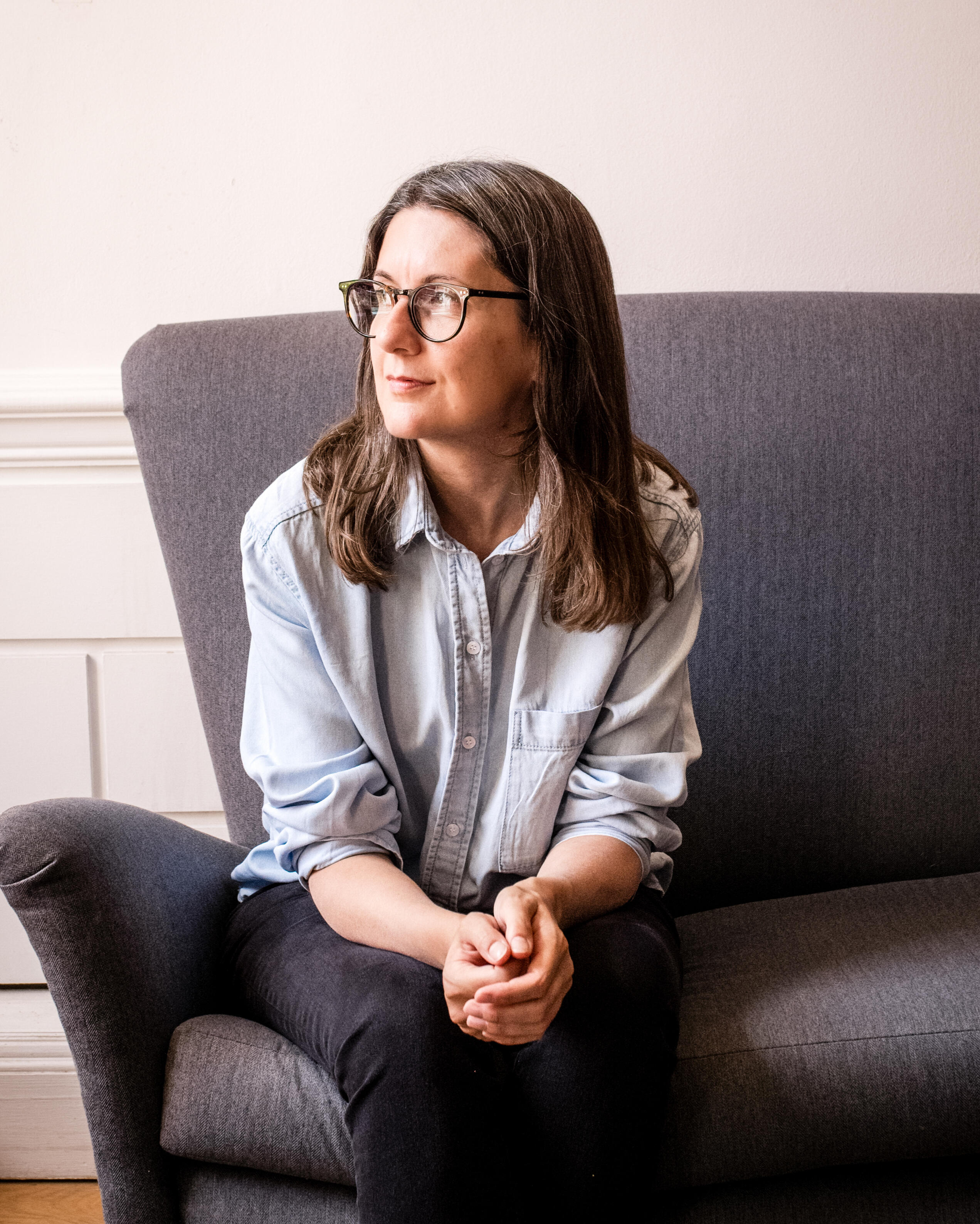 A woman (Jess) sitting on a sofa, looking towards the window with her hands resting together in her lap.